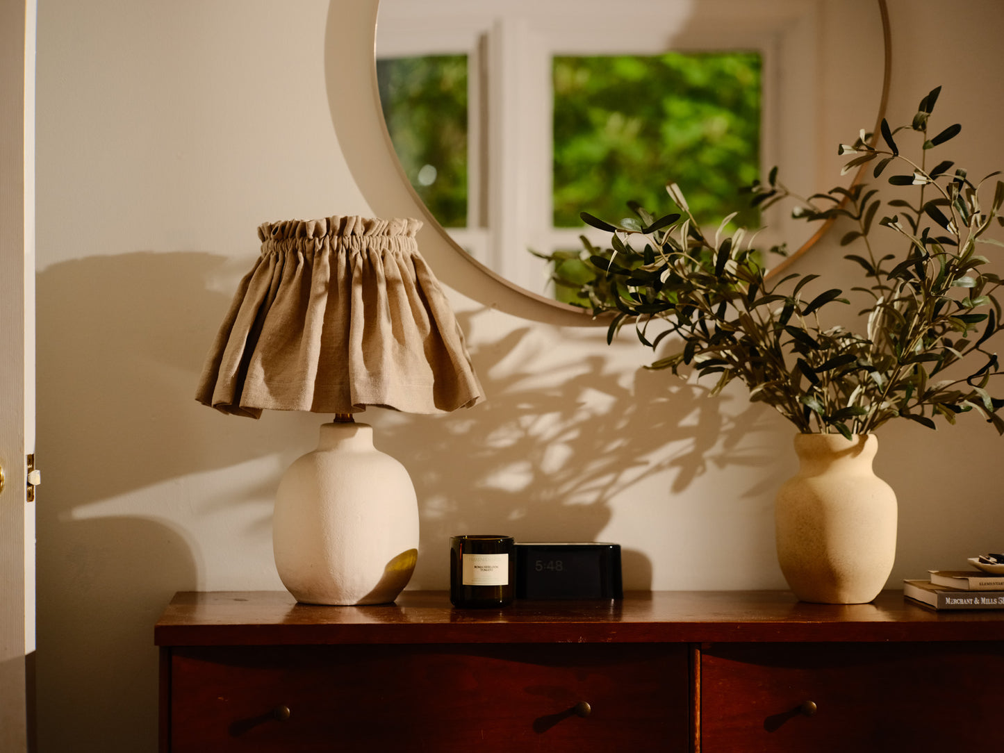 Lamp with a beige shade on a wooden surface next to a plant and a mirror reflecting greenery.