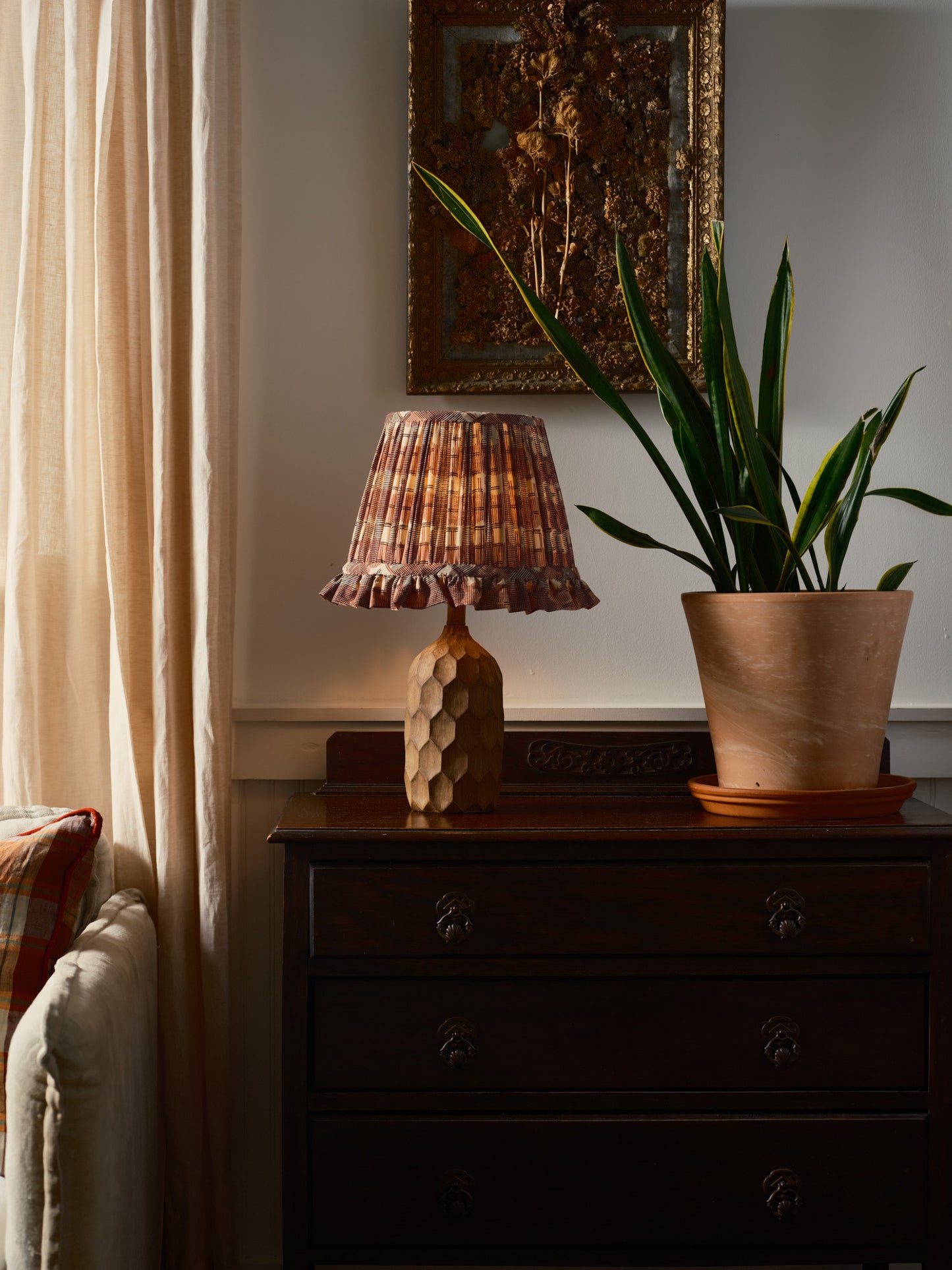Lamp with plaid shade on a wooden side table next to a plant, with a painting in the background.
