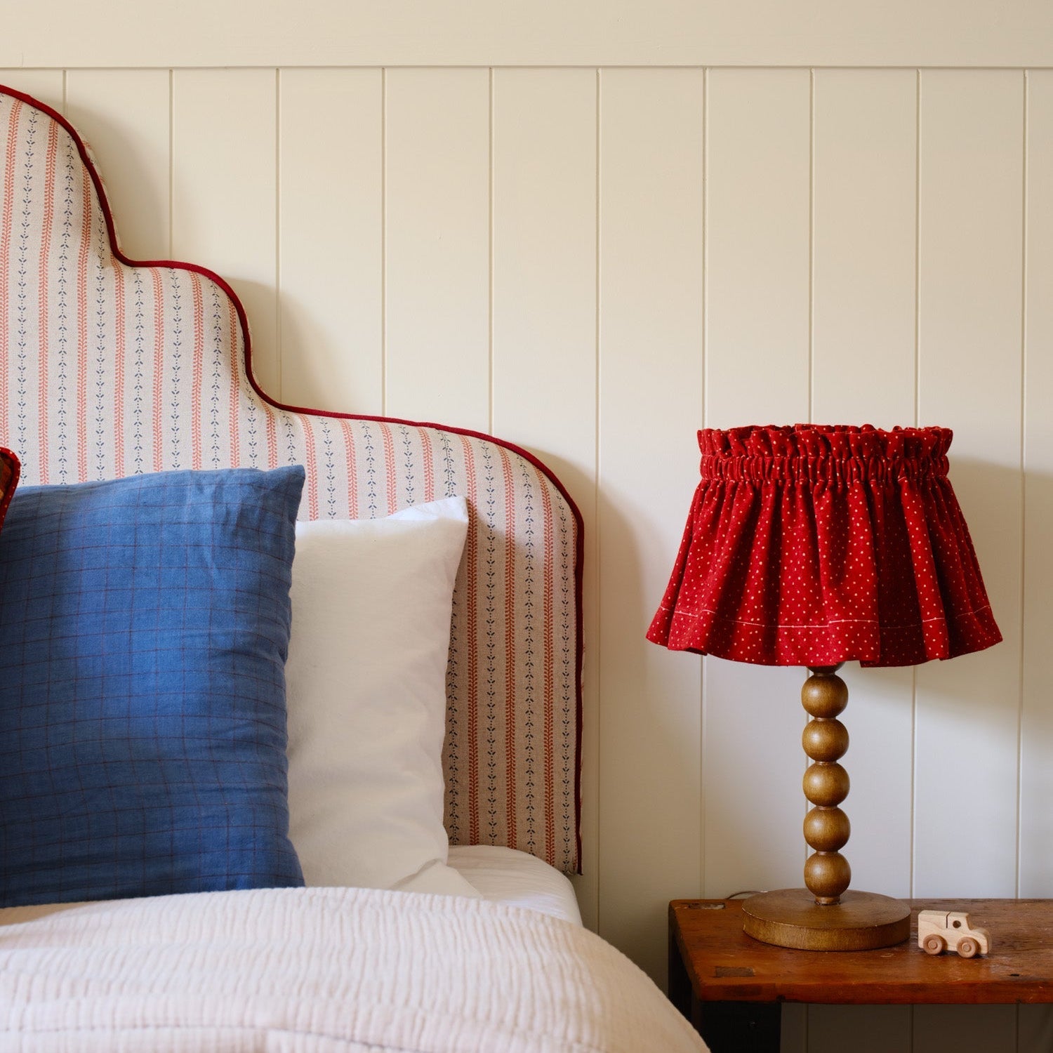 red lampshade on a nightstand next do a stripped headboard and decorative pillows. 