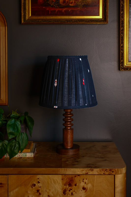 Black pleated lampshade with red accents on a wooden lamp base against a dark wall.