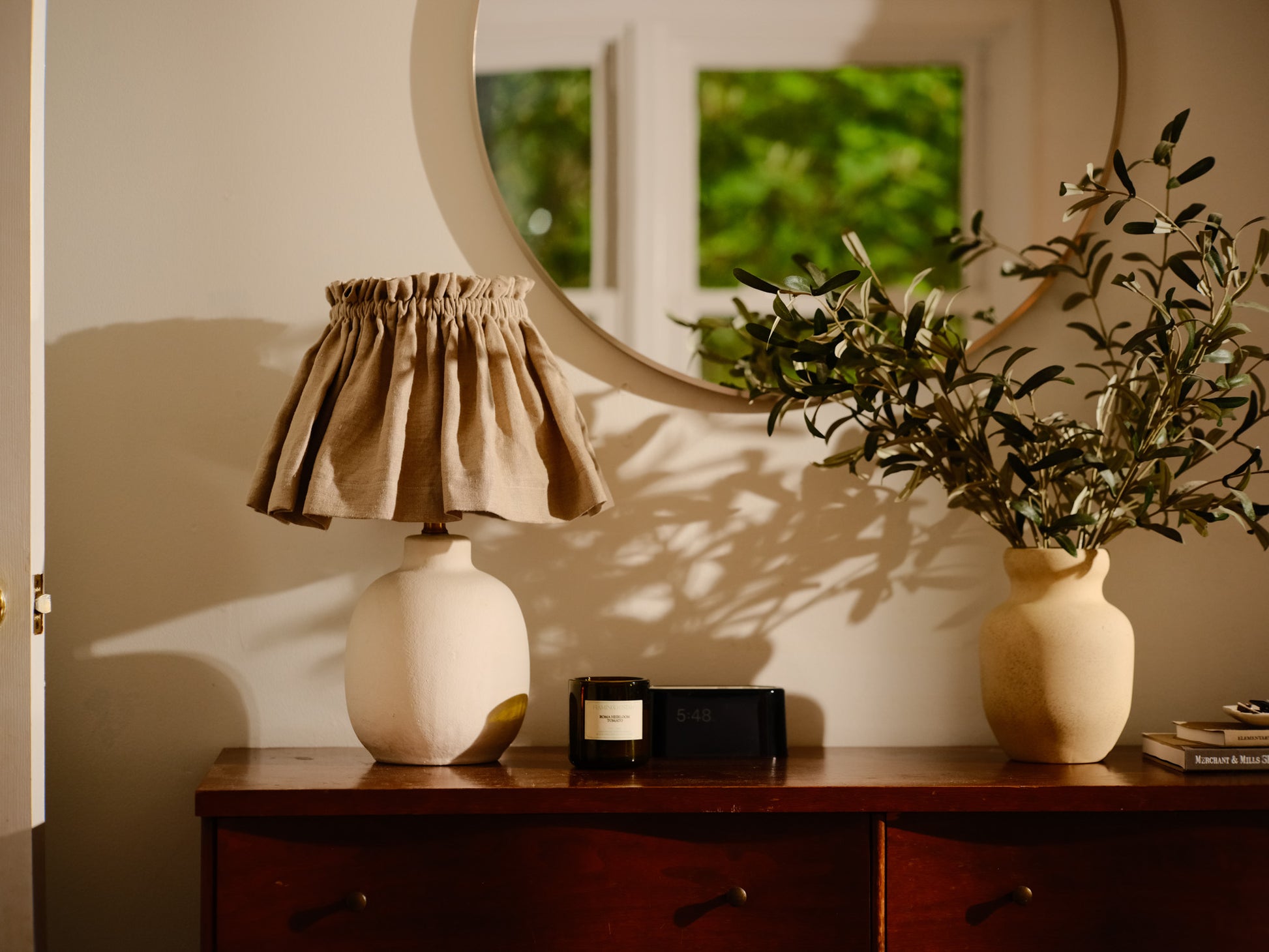 Lamp with a beige shade on a wooden surface next to a plant and a mirror reflecting greenery.