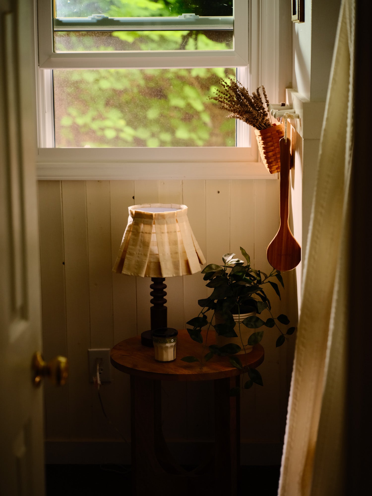 Small round wooden table with a lamp and plant against a window with a view of greenery.
