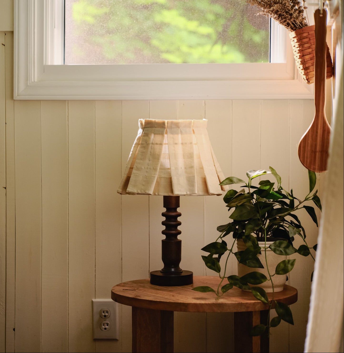 Small round wooden table with a lamp and plant against a wall with a window.