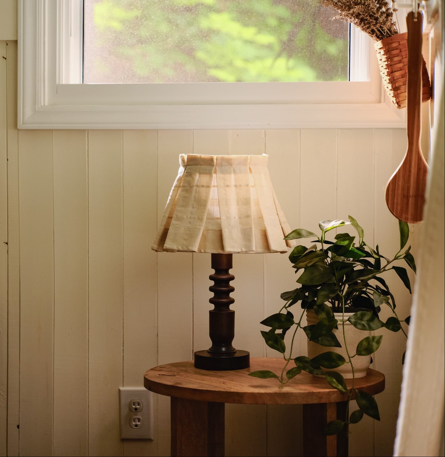 Small round wooden table with a lamp and plant against a wall with a window.