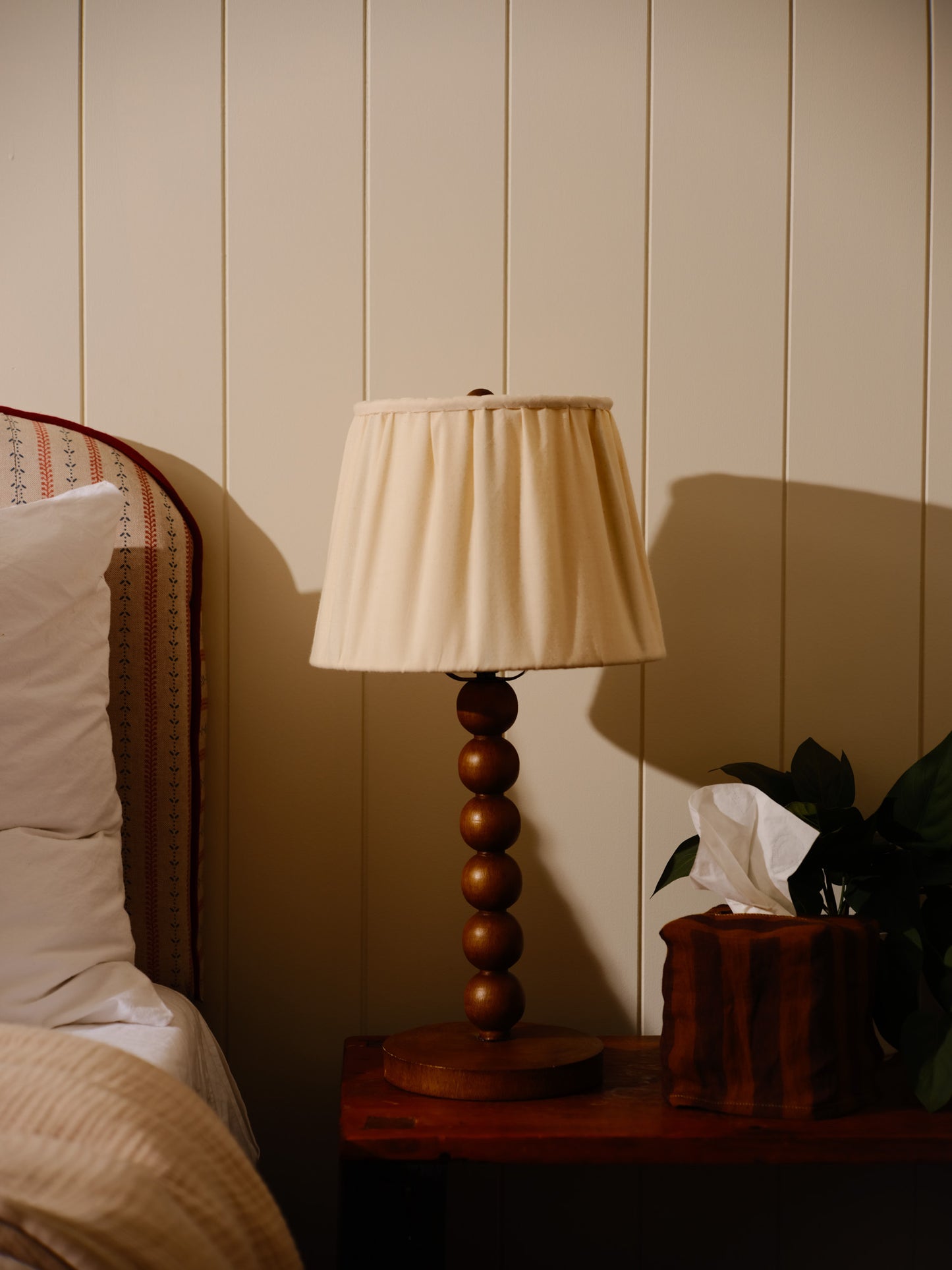 Table lamp with wooden base and beige shade on a bedside table against a wall.