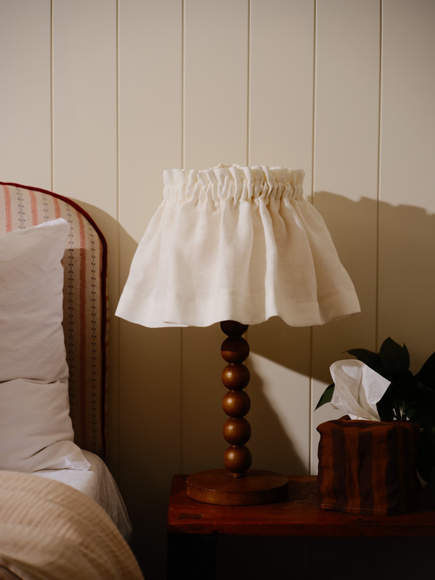 Lamp with a white lampshade on a wooden table next to a bed with striped headboard.