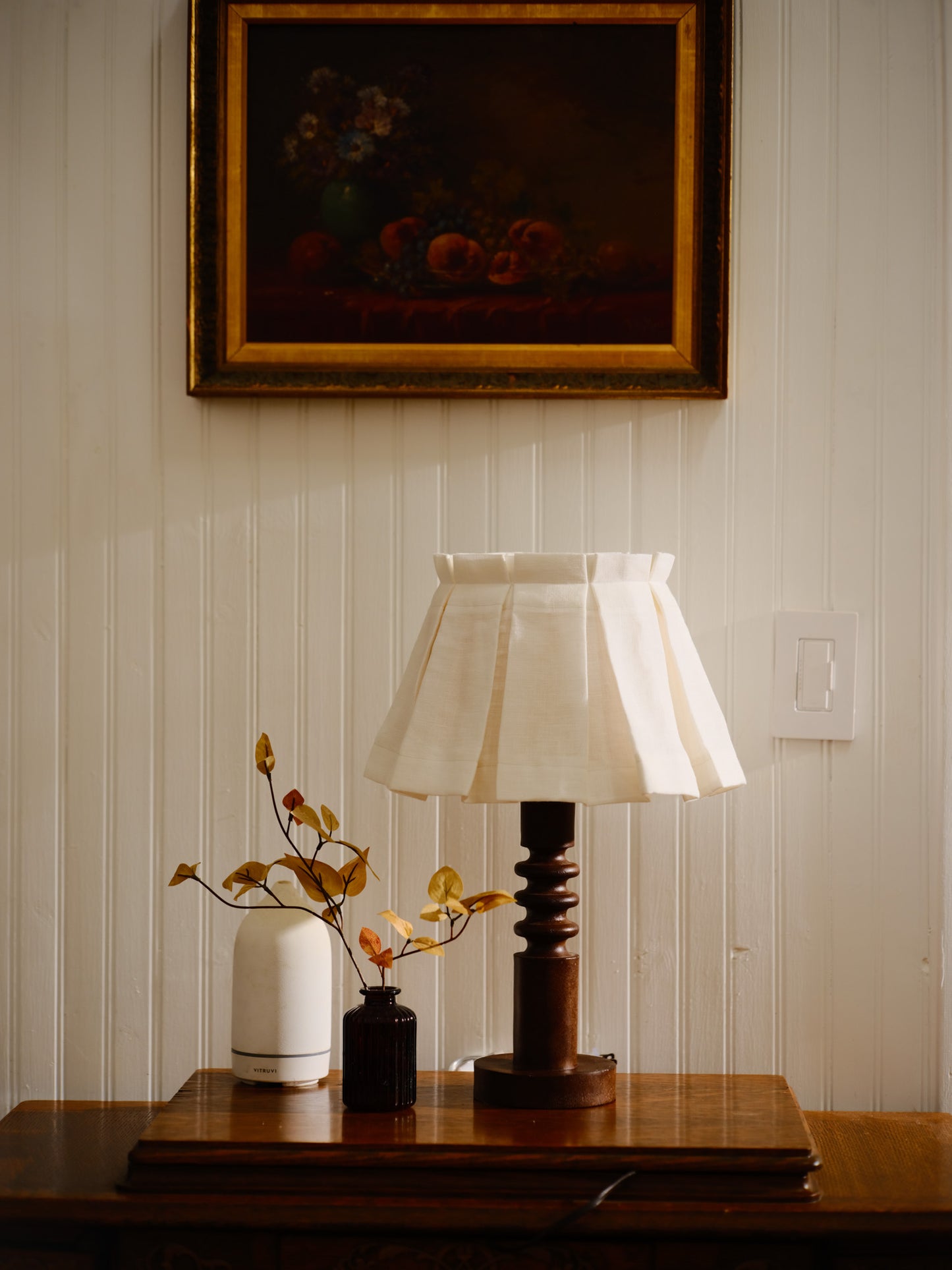Table lamp with white shade on a wooden surface, with a painting in the background.