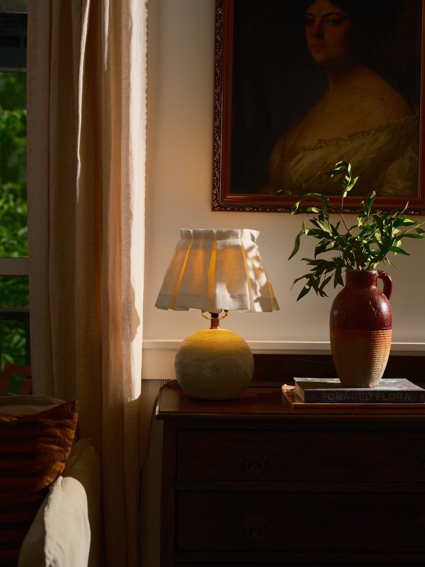 Lamp with a white box pleated shade on a wooden surface with a  plant in the background