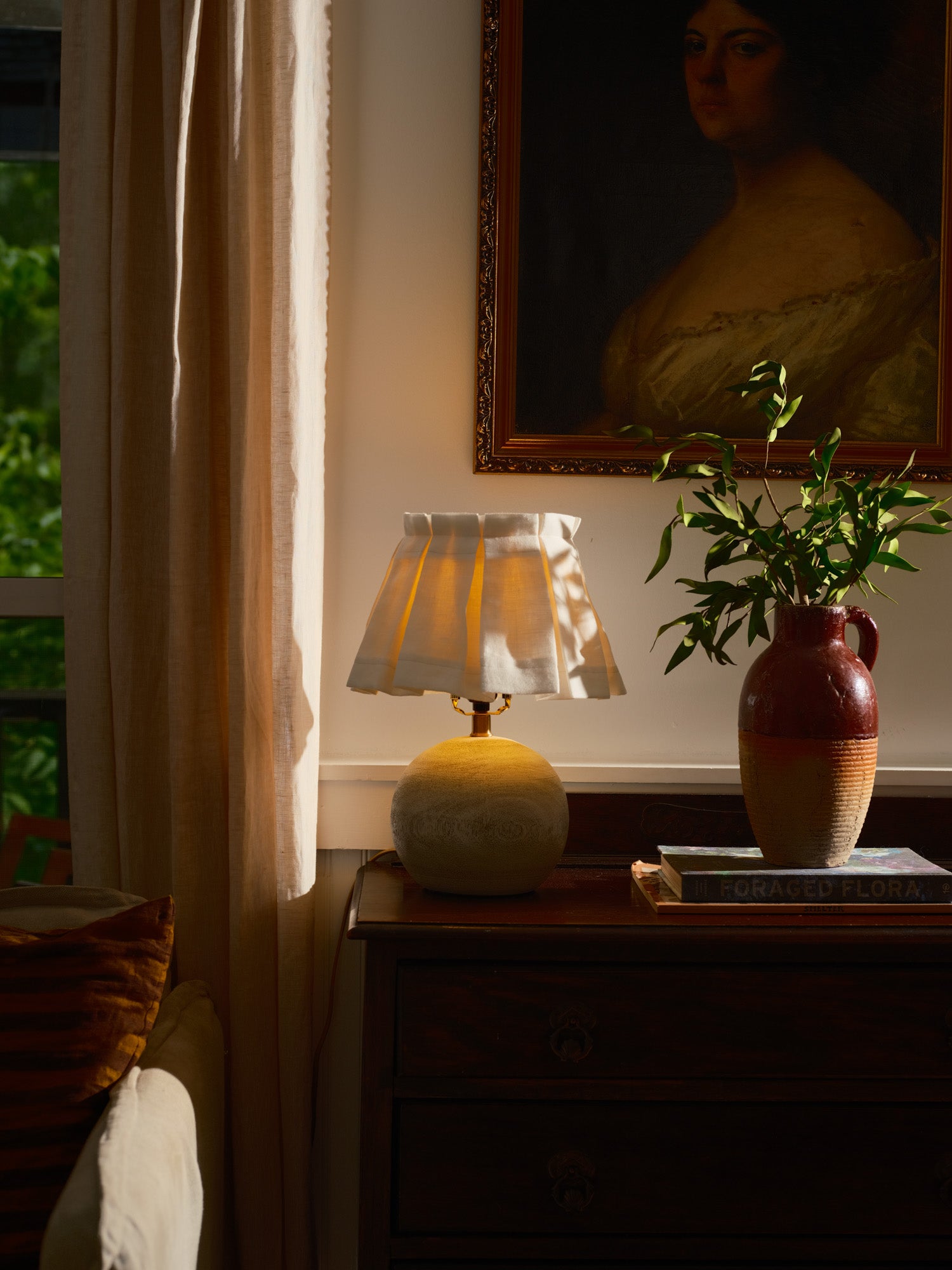 Lamp with a white box pleated shade on a wooden surface with a  plant in the background
