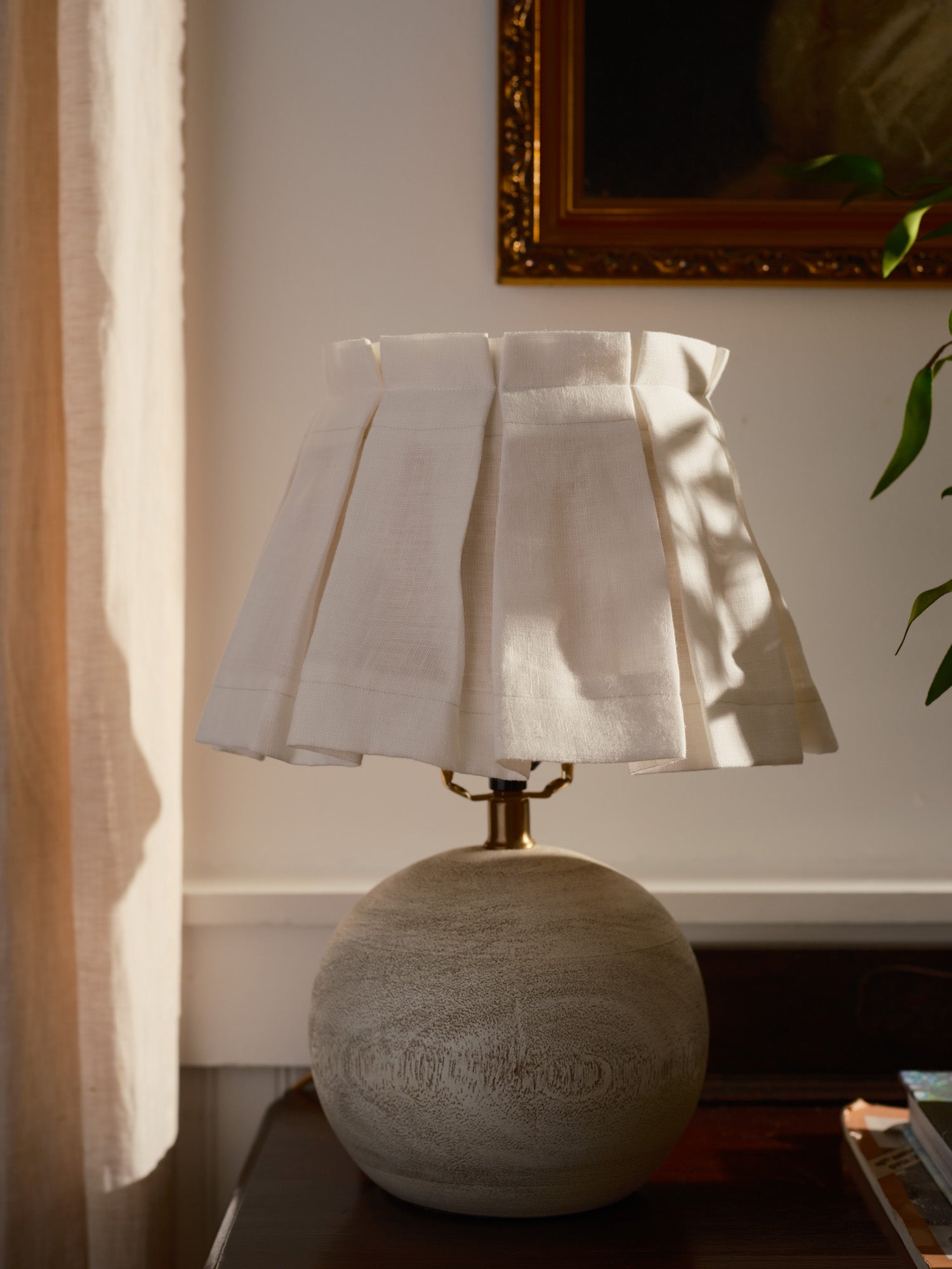 Decorative lamp with a white lampshade on a wooden surface, with a mirror and plant in the background.