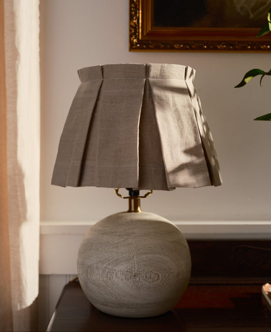 Decorative lamp with pleated shade on a wooden surface, with a painting in the background.