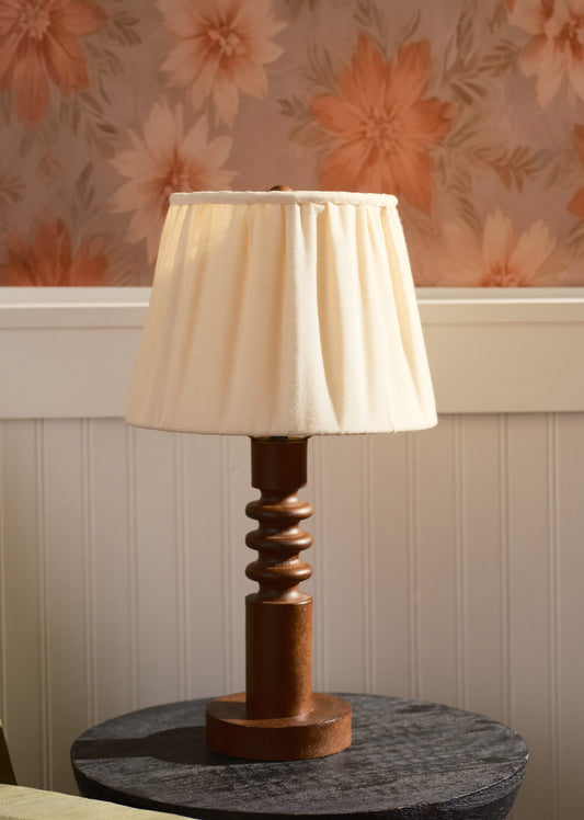 Table lamp with a wooden base and white shade on a round table against floral wallpaper.