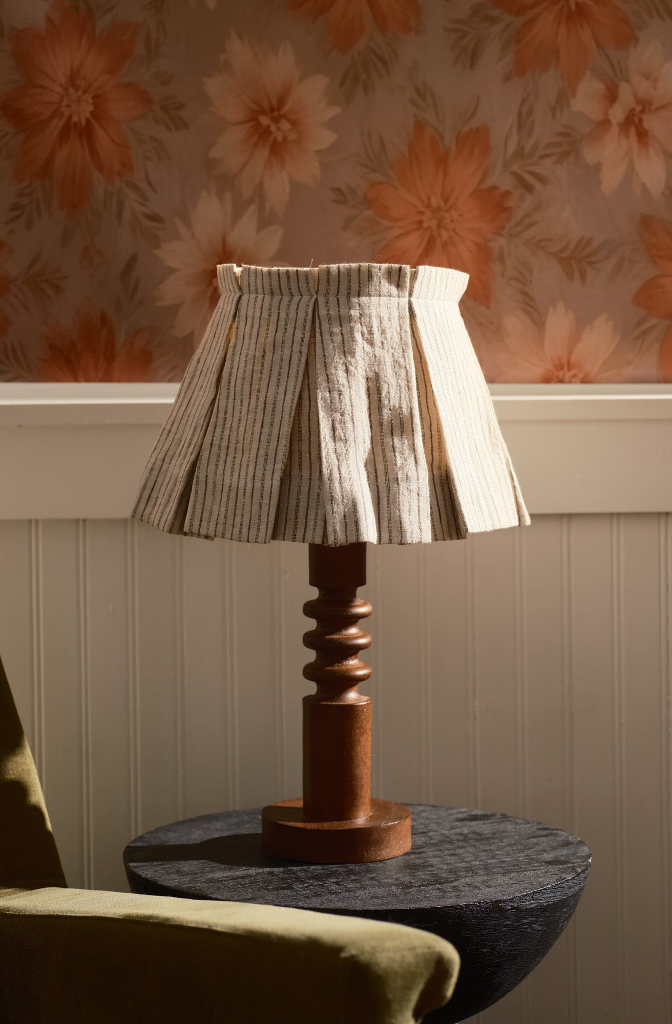 Table lamp with box pleated shade on a round table against floral wallpaper.