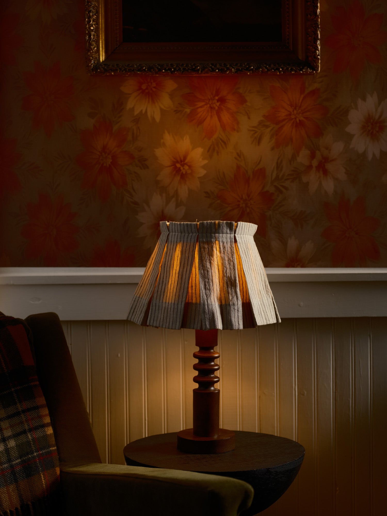 Lamp with a stripped shade on a table against a floral wallpapered wall.