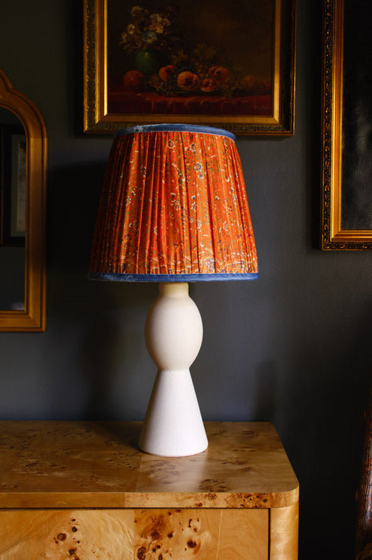 White lamp with an orange pleated shade on a wooden table against a dark wall with framed artwork.