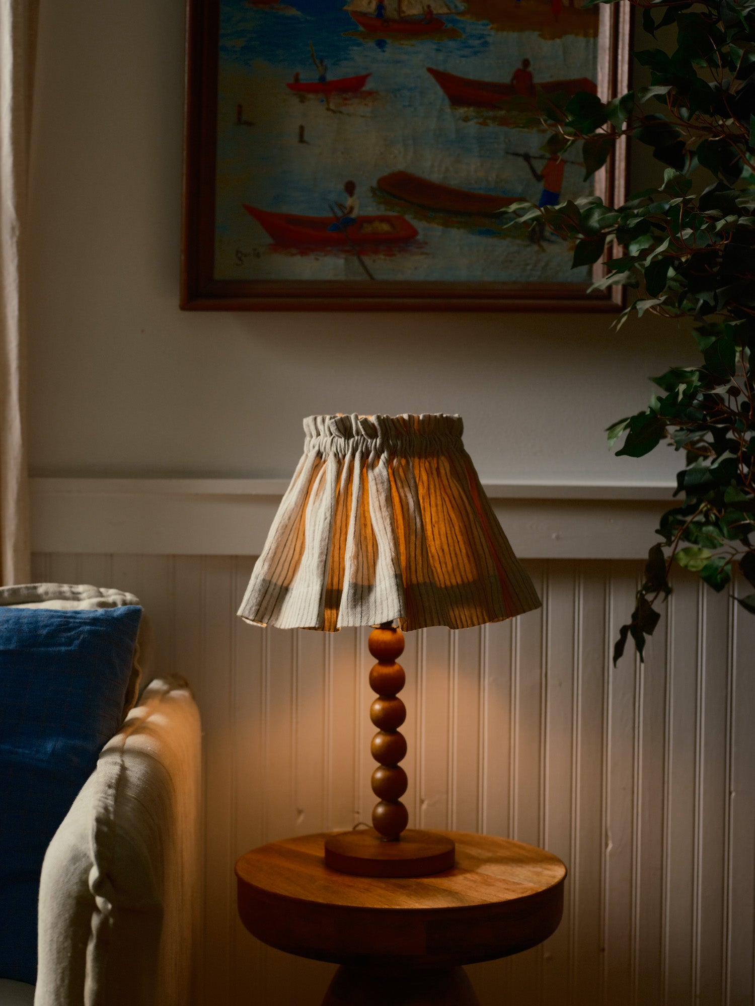 Wooden lamp with striped shade on a side table in a room with a painting of boats on the wall.