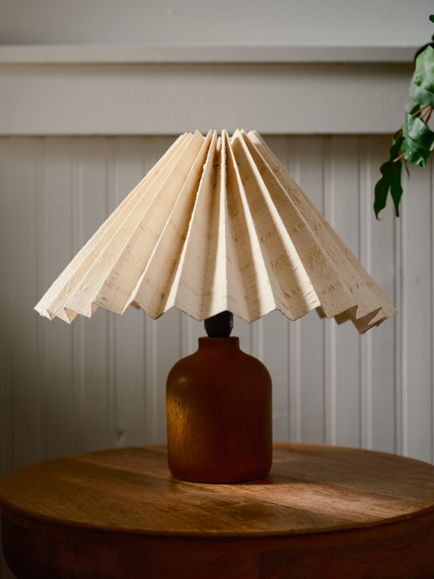 Lamp with a pleated beige shade on a wooden table against a white paneled wall.